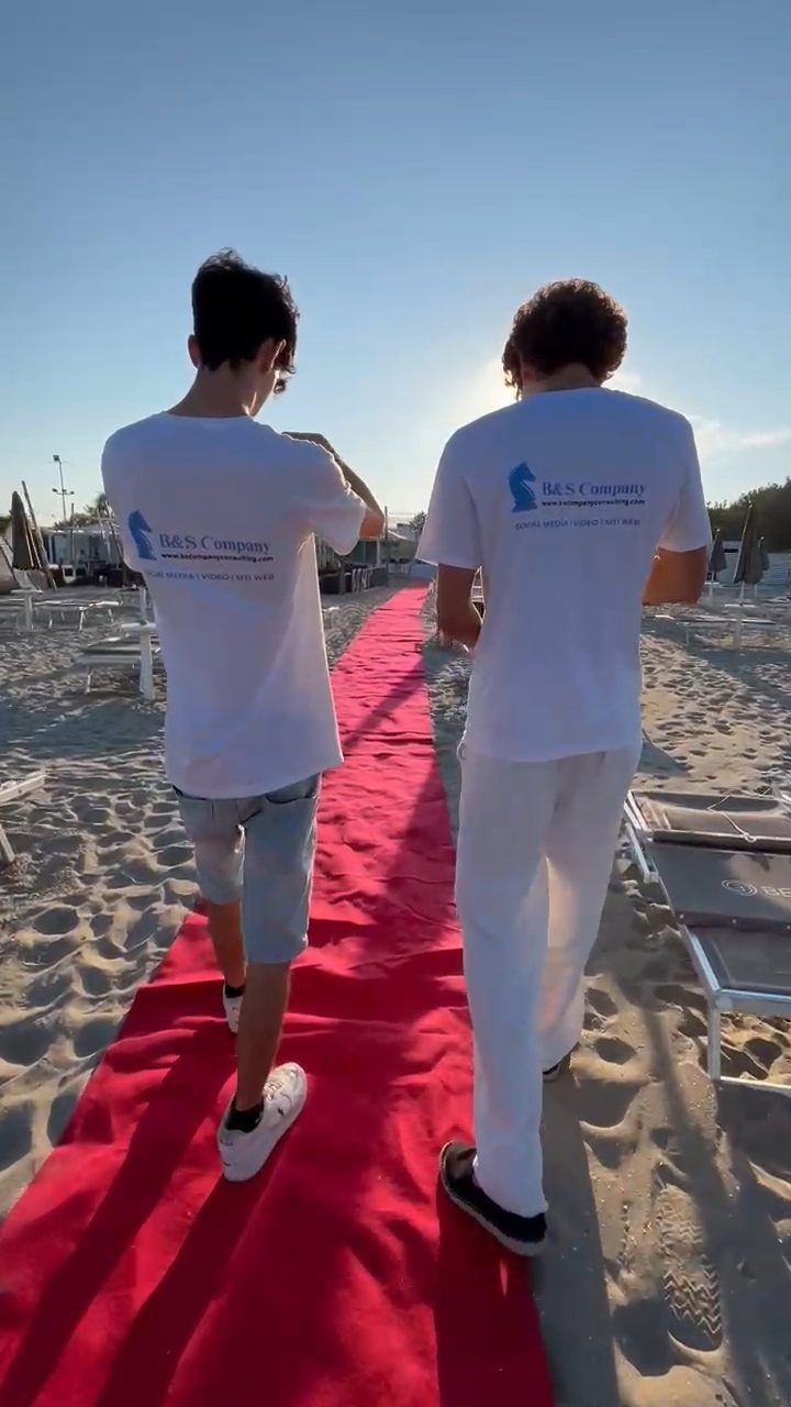 Due operatori di B&S Company camminano sul tappeto rosso di un evento in spiaggia al tramonto